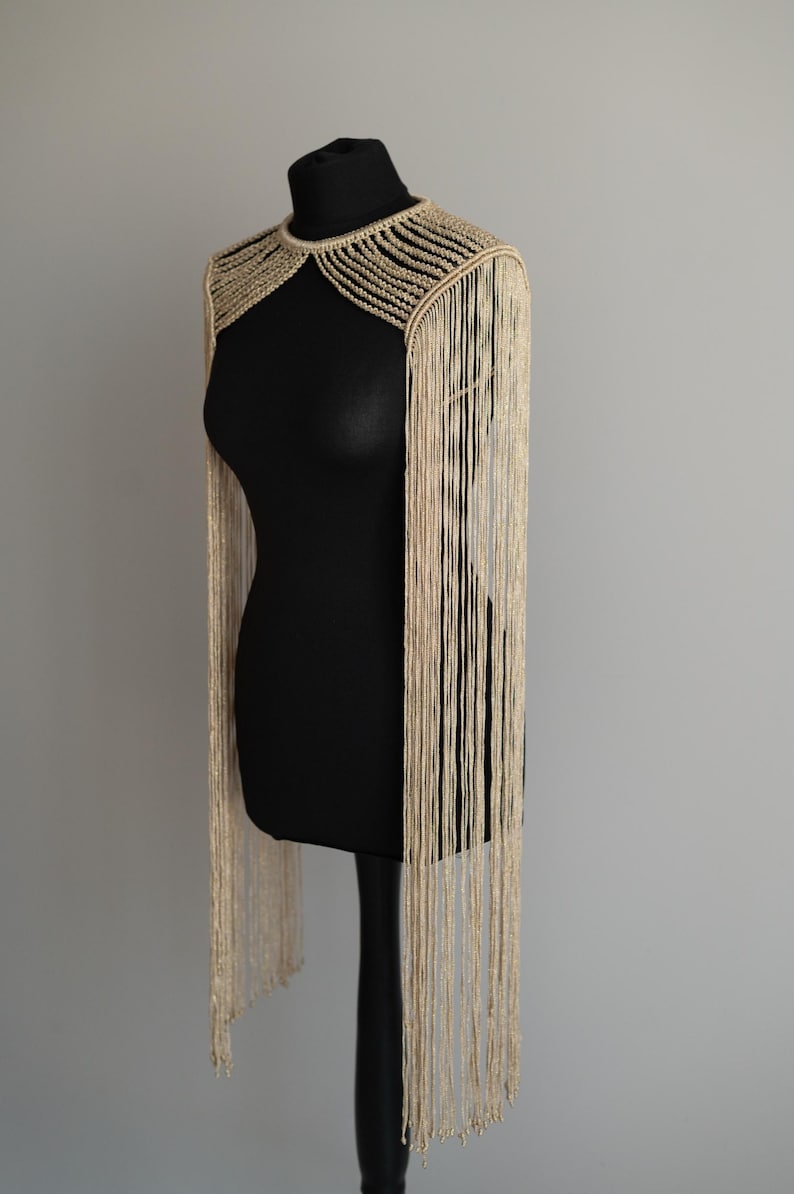 Peut inclure: Une pi&egrave;ce d'&eacute;paule en macram&eacute; beige avec un col montant et des d&eacute;tails nou&eacute;s complexes autour du cou. De longs fils de franges fins et pendants s'&eacute;tendent des &eacute;paules jusqu'&agrave; la taille, cr&eacute;ant un aspect textur&eacute; et superpos&eacute;.