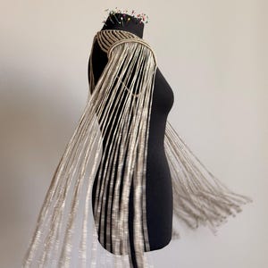 Peut inclure: Une pi&egrave;ce d'&eacute;paule en macram&eacute; beige avec de longs brins de franges en cascade qui tombent des &eacute;paules. Le v&ecirc;tement est construit avec des motifs de cordons nou&eacute;s complexes et est pr&eacute;sent&eacute; sur un buste de mannequin noir avec des &eacute;pingles au cou.