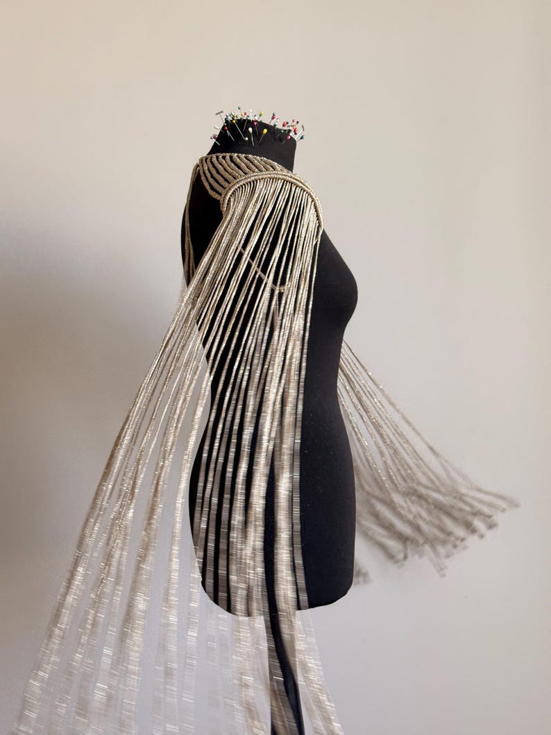 Peut inclure: Une pi&egrave;ce d'&eacute;paule en macram&eacute; beige avec de longs brins de franges en cascade qui tombent des &eacute;paules. Le v&ecirc;tement est construit avec des motifs de cordons nou&eacute;s complexes et est pr&eacute;sent&eacute; sur un buste de mannequin noir avec des &eacute;pingles au cou.