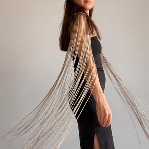 Peut inclure: Une robe bustier noire dot&eacute;e de longues franges m&eacute;talliques argent&eacute;es sur les &eacute;paules qui tombent le long des bras. Le v&ecirc;tement est une pi&egrave;ce noire unie et ajust&eacute;e avec une fente lat&eacute;rale, rehauss&eacute;e par des cha&icirc;nes argent&eacute;es scintillantes.