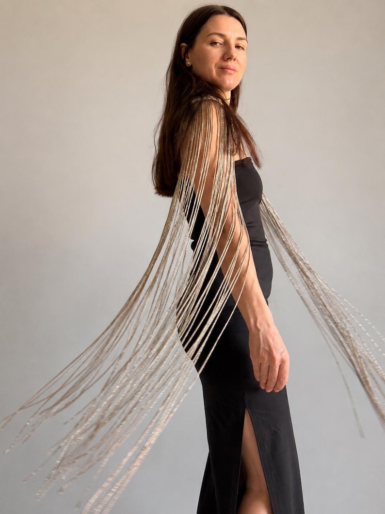 Peut inclure: Une robe bustier noire dot&eacute;e de longues franges m&eacute;talliques argent&eacute;es sur les &eacute;paules qui tombent le long des bras. Le v&ecirc;tement est une pi&egrave;ce noire unie et ajust&eacute;e avec une fente lat&eacute;rale, rehauss&eacute;e par des cha&icirc;nes argent&eacute;es scintillantes.