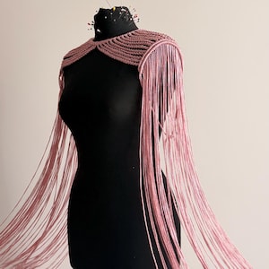 Peut inclure: Une pi&egrave;ce d'&eacute;paule en macram&eacute; rose poudr&eacute; pr&eacute;sentant un design structur&eacute; &agrave; n&oelig;uds qui se transforme en longs brins de franges fluides descendant sous la taille, pr&eacute;sent&eacute;e sur un buste de mannequin noir avec plusieurs &eacute;pingles &agrave; couture.