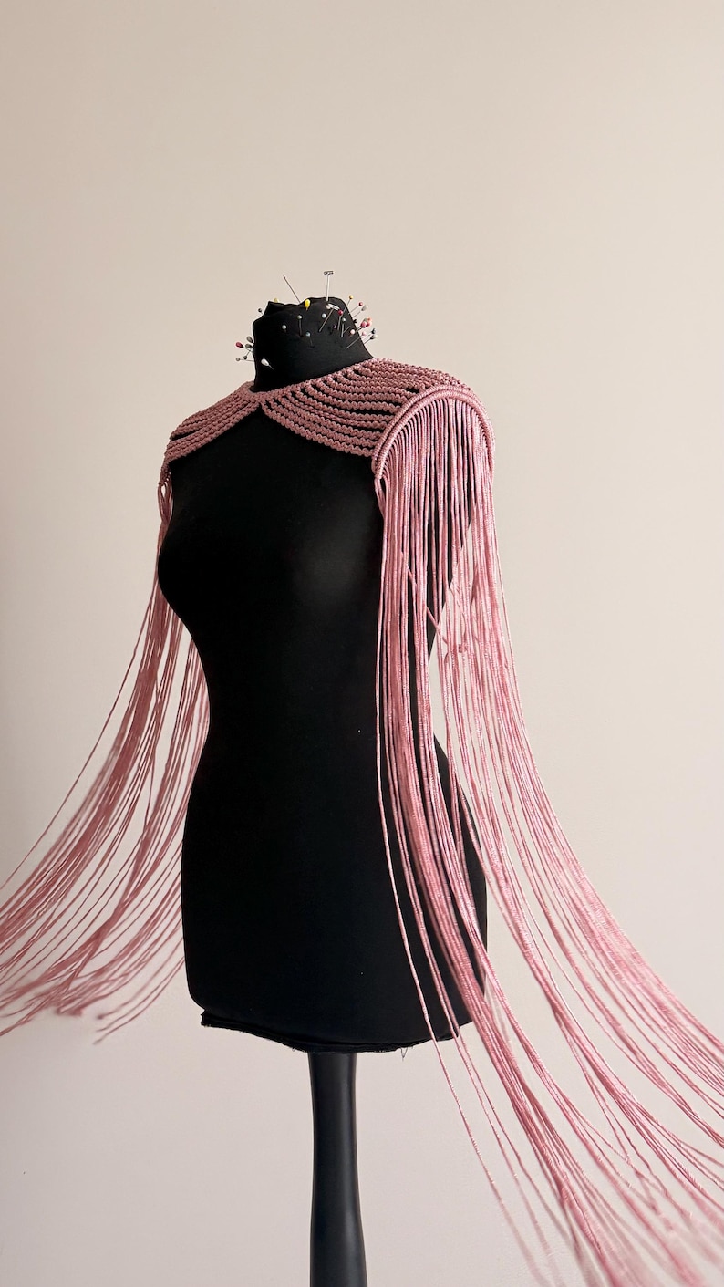 Peut inclure: Une pi&egrave;ce d'&eacute;paule en macram&eacute; rose poudr&eacute; pr&eacute;sentant un design structur&eacute; &agrave; n&oelig;uds qui se transforme en longs brins de franges fluides descendant sous la taille, pr&eacute;sent&eacute;e sur un buste de mannequin noir avec plusieurs &eacute;pingles &agrave; couture.
