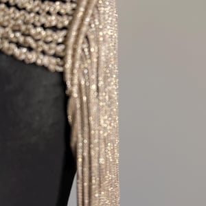 Peut inclure: Un v&ecirc;tement compos&eacute; d'une base noire avec une superposition de franges dor&eacute;es m&eacute;talliques. Les franges sont constitu&eacute;es de nombreuses m&egrave;ches fines et scintillantes qui pendent verticalement depuis l'&eacute;paule, cr&eacute;ant un aspect textur&eacute;.