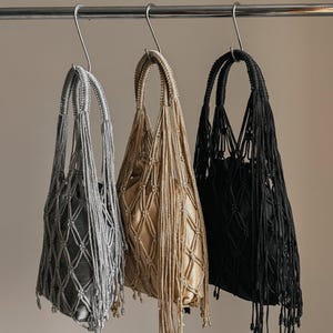 May include: Three macrame tote bags in silver, gold, and black hang from a metal bar. Each bag features a knotted net design with a drawstring closure and long fringe. The bags are suspended against a neutral background.