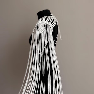 Può includere: Stola a spalla in macramè bianco con design geometrico e lunghe frange. Il tessuto fatto a mano presenta nodi e drappeggi intricati, creando un accessorio in stile bohémien. La stola è esposta su un manichino nero.