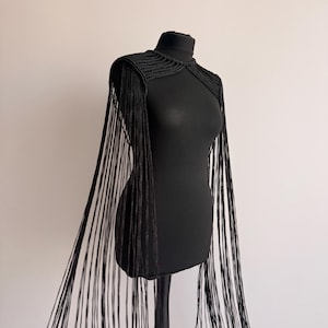 Könnte beinhalten: Ein schwarzes gehäkeltes Schulterstück mit hohem Ausschnitt und langen, fließenden Fransen, die von den Schultern bis zum Saum reichen. Das Kleidungsstück ist mit einem strukturierten Rippenstrickmuster an Brust und Schultern gefertigt.