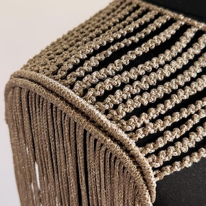 Peut inclure: Un accessoire d'&eacute;paule dor&eacute; m&eacute;tallique compos&eacute; d'une s&eacute;rie de cordons tress&eacute;s horizontaux qui se prolongent en longues franges fluides. La pi&egrave;ce pr&eacute;sente un bord incurv&eacute; et structur&eacute; qui repose sur l'&eacute;paule, avec une finition scintillante et textur&eacute;e sur l'ensemble du design.