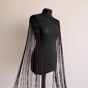 Peut inclure: Une pi&egrave;ce d'&eacute;paule crochet&eacute;e noire dot&eacute;e d'un col haut et de longues franges fluides qui descendent des &eacute;paules jusqu'&agrave; l'ourlet. Le v&ecirc;tement est r&eacute;alis&eacute; avec un motif de points c&ocirc;tel&eacute;s textur&eacute;s sur la partie sup&eacute;rieure de la poitrine et des &eacute;paules.