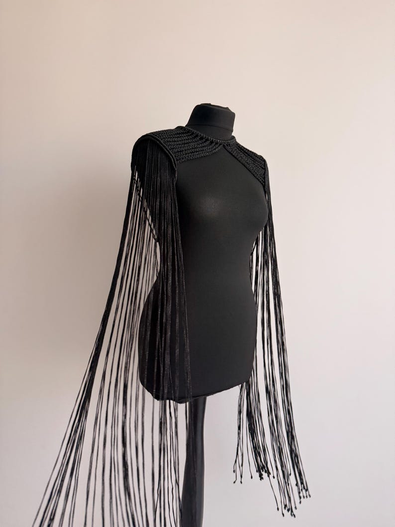 Peut inclure: Une pi&egrave;ce d'&eacute;paule crochet&eacute;e noire dot&eacute;e d'un col haut et de longues franges fluides qui descendent des &eacute;paules jusqu'&agrave; l'ourlet. Le v&ecirc;tement est r&eacute;alis&eacute; avec un motif de points c&ocirc;tel&eacute;s textur&eacute;s sur la partie sup&eacute;rieure de la poitrine et des &eacute;paules.