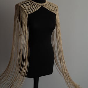 May include: A gold-colored, fringed shoulder wrap displayed on a black mannequin. The wrap features a beaded collar and long, flowing fringe that extends down the arms. The garment is designed for formal wear.
