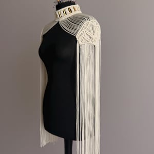 Op de afbeelding: Crèmekleurige macramé schouderwikkel met franjedetails. De wikkel heeft een hoge kraag en geometrische patronen op de schouders. Het kledingstuk wordt gepresenteerd op een zwarte mannequin tegen een grijze achtergrond.