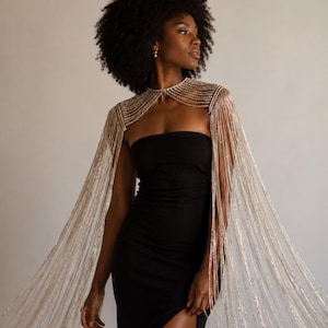May include: A black strapless floor-length dress with a high side slit, paired with a silver metallic chain fringe capelet that drapes over the shoulders and extends down the arms, featuring multiple layered strands of shimmering silver chains.