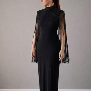 Puede incluir: Un vestido negro largo hasta el suelo con cuello alto y flecos en los hombros y las mangas. El vestido es entallado y está hecho de una tela suave y fluida. Los flecos añaden un toque dramático al diseño elegante.