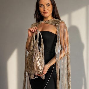 May include: A gold beaded cape with long fringe and a matching handbag. The cape is draped over a black dress. The handbag has a quilted pattern and a beaded handle. The overall look is elegant and glamorous.
