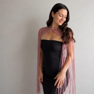 May include: A dusty rose macrame shoulder wrap featuring a structured, woven neckline and long, flowing fringe that extends down the arms. The wrap is worn over a simple, solid black strapless dress, creating a contrast between the textured yarn.