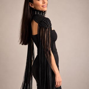May include: A black macrame shoulder piece featuring a high neck collar with silver-toned hardware accents. The garment includes intricate knotted patterns on the shoulders and long, flowing black fringe that extends down the arms and back.