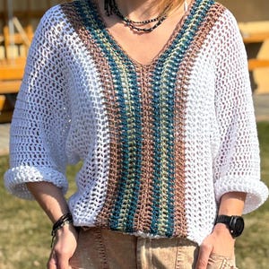 Puede incluir: Top de crochet blanco con cuello en V y mangas tres cuartos. El panel central presenta rayas verticales en marrón, verde azulado y amarillo. Las mangas están dobladas. Se ve un reloj y una pulsera negros.