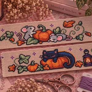 Puede incluir: Dos marcapáginas rectangulares de punto de cruz sobre tela beige. El superior muestra dos ratones grises con una calabaza y hojas de otoño. El inferior muestra un gato negro descansando sobre una calabaza junto a un farol y hiedra.