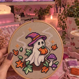 Könnte beinhalten: Ein Kreuzstich-Stickprojekt in einem runden Holzrahmen, das einen weißen Geist mit einem violetten Hexenhut zeigt, der einen orangefarbenen Kürbis hält, umgeben von herbstlichen Blättern und violetten Pilzen auf beigem Stoff.