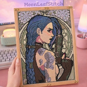 Op de afbeelding: Kruissteekkunstwerk van een personage met blauw haar en een tatoeage, die een wapen vasthoudt. Het kunstwerk is ingelijst in een licht houten frame met goudkleurige hoekaccenten. De tekst "MoonLeafStitch" is zichtbaar.