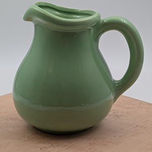 May include: A light green ceramic pitcher with a smooth, glossy finish. The vessel features a rounded, bulbous body, a curved handle on the right side, and a distinct pouring spout at the top rim. The surface reflects light evenly across its curves.