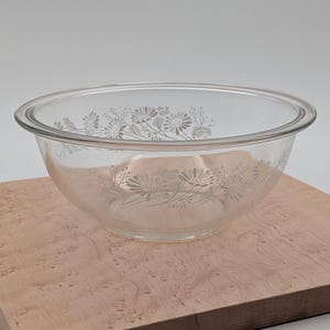 Puede incluir: Un cuenco de vidrio transparente con un diseño floral. El cuenco tiene un borde ancho y una forma redondeada. El diseño floral está grabado en el vidrio y presenta un patrón repetido de flores y hojas. El cuenco está sobre una superficie de madera.