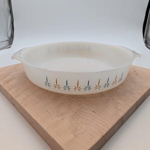 May include: An oval, white glass casserole dish with a decorative pattern of blue and orange starbursts. The dish has two handles and is set on a light-colored wooden cutting board. A vintage kitchenware item.