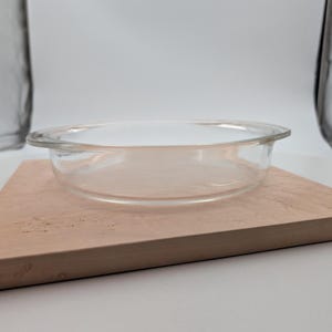 Puede incluir: Un recipiente para hornear redondo de vidrio transparente con un borde ligeramente ensanchado. El recipiente está sobre una tabla de cortar de madera marrón claro. El vidrio es transparente.