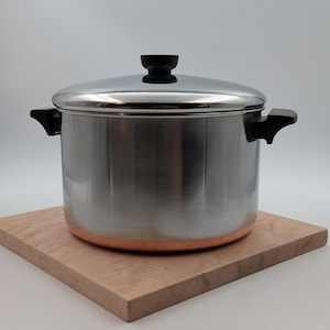 May include: A stainless steel stockpot with a lid and black handles. The pot has a copper-colored bottom and sits on a light-colored wooden cutting board. The lid has a black handle. The pot is designed for cooking.