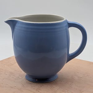 May include: A periwinkle blue ceramic pitcher with a rounded body, a curved handle, and a wide pouring spout. The exterior features subtle horizontal ridges near the top rim, while the interior is finished in a smooth, off-white glaze.