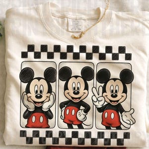 May include: Cream-colored sweatshirt featuring a graphic of Mickey Mouse in three different poses. Mickey is wearing red shorts, black gloves, and shoes. The design is framed by a black and white checkered pattern. A gold necklace is draped across the shirt.