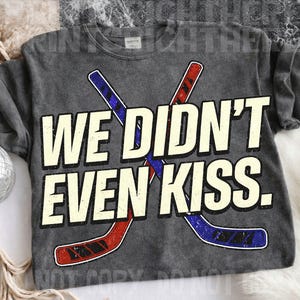 May include: Dark gray sweatshirt with the text "WE DIDN'T EVEN KISS." The design includes two crossed hockey sticks in red and blue. The sweatshirt is a casual, comfortable style.