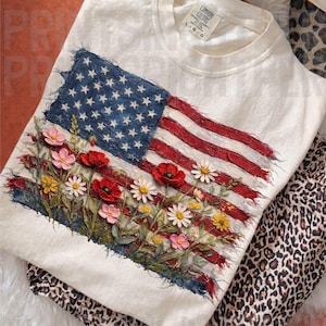 Op de afbeelding: Crèmekleurig T-shirt met een versleten Amerikaans vlagontwerp met bloemaccenten. De vlag bevat rode en witte strepen, een blauw veld met sterren en een kleurrijk scala aan bloemen. Gecombineerd met een broek met luipaardprint.