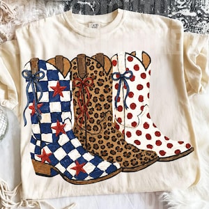 Op de afbeelding: Crèmekleurig T-shirt met een afbeelding van vier cowboylaarzen. De laarzen tonen verschillende ontwerpen: blauw-wit geruit, luipaardprint en wit met rode stippen. Rode ster- en strikaccenten zijn toegevoegd.