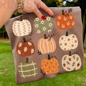 Puede incluir: Una camiseta marrón con un patrón repetitivo de nueve calabazas ilustradas en varios colores, incluyendo naranja, crema y verde salvia. Cada calabaza muestra detalles decorativos únicos como motivos florales, hojas y cuadros.