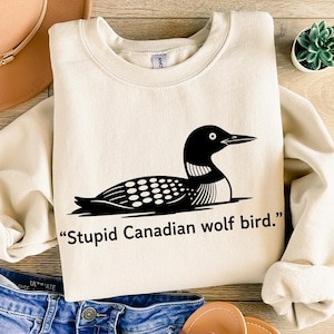 Pode incluir: Camisola creme com um gráfico preto de um mergulhão e o texto "Stupid Canadian wolf bird." O mergulhão é retratado de forma minimalista, com cabeça e corpo pretos e manchas brancas. A camisola é de estilo casual.