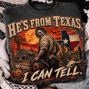 Puede incluir: Una sudadera gris oscuro con un gr&aacute;fico de un personaje sosteniendo una motosierra en un entorno rural con un molino, un granero, una calabaza, un cactus y la bandera de Texas. El texto dice HE'S FROM TEXAS, I CAN TELL en una fuente desgastada.