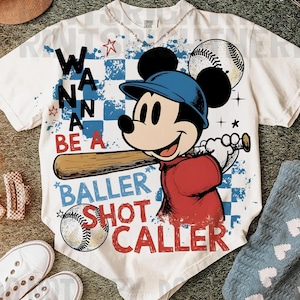 May include: White t-shirt featuring Mickey Mouse in a baseball uniform, holding a bat. The shirt has the text "Wanna Be A Baller Shot Caller" with baseball and star graphics. The design is on a checkered blue and white background.