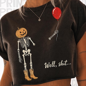 Op de afbeelding: Een donker antracietgrijs t-shirt met korte mouwen en een afbeelding van een skelet met een pompoenhoofd en bruine cowboylaarzen. Een losse skeletarm houdt een rode ballon vast. De tekst "Well, shit..." is in het wit gedrukt.