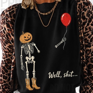 Op de afbeelding: Een zwart shirt met lange mouwen en een luipaardprint op de mouwen. Op de voorkant staat een skelet met een pompoenhoofd, bruine cowboylaarzen en een rode ballon in de hand. De tekst luidt, Well, shit...