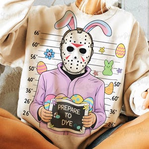 Puede incluir: Una sudadera color crema con un gráfico humorístico. El diseño incluye una máscara de hockey con orejas de conejo, una camisa rosa y un letrero que dice "PREPARE TO DYE". También hay huevos de Pascua y flores.