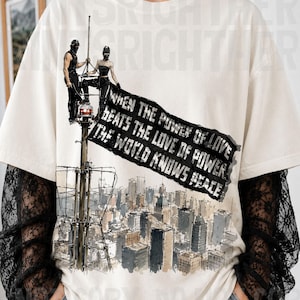 May include: An off-white short-sleeve t-shirt with black lace long sleeves. The graphic features two figures on a tall antenna tower holding a black banner with the text: WHEN THE POWER OF LOVE BEATS THE LOVE OF POWER THE WORLD KNOWS PEACE.