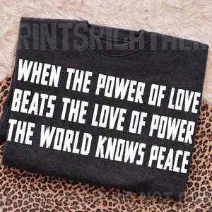 May include: A charcoal gray crewneck sweatshirt featuring white block lettering that reads: WHEN THE POWER OF LOVE BEATS THE LOVE OF POWER THE WORLD KNOWS PEACE. The garment has a distressed, vintage-inspired wash and a classic long-sleeve design.