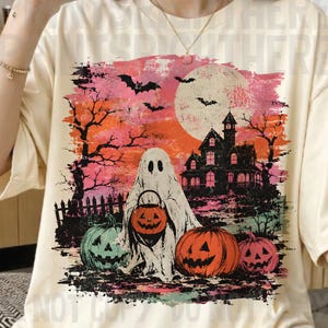 May include: A cream-colored t-shirt featuring a graphic print of a ghost holding a jack-o'-lantern bucket, a haunted house, bats, a full moon, and additional pumpkins against a vibrant orange and pink sunset background with black silhouettes.