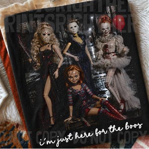 May include: A black t-shirt featuring a graphic print of four horror-themed dolls. The dolls are dressed in dark attire, with one holding a red balloon. The text "i'm just here for the boos" is printed in white script at the bottom of the graphic.