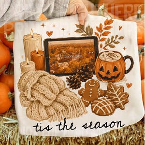 May include: A white fabric featuring a graphic print of a tablet displaying a town scene with the text Gilmore girls, surrounded by three candles, pinecones, a pumpkin-shaped mug, a gingerbread man, a cookie, and a knit scarf with the text tis the season.
