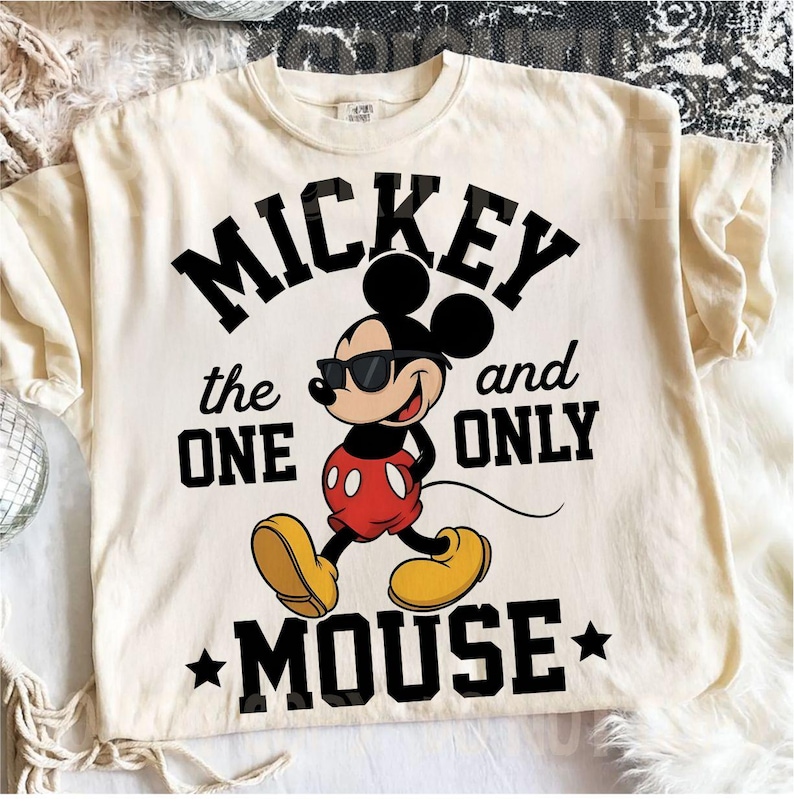 May include: Cream-coloured t-shirt with a graphic of Mickey Mouse wearing sunglasses, and the text "MICKEY the ONE and ONLY MOUSE" in black. The design includes stars and a classic cartoon style.