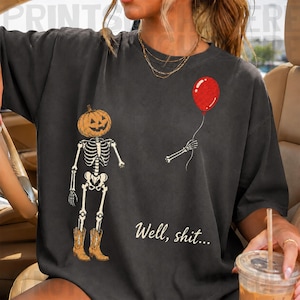 Op de afbeelding: Een antracietgrijs t-shirt met een afbeelding van een skelet met een pompoenhoofd en bruine cowboylaarzen. Het skelet houdt een rode ballon vast in &eacute;&eacute;n hand. De tekst "Well, shit..." staat in een wit cursief lettertype onder de afbeelding.
