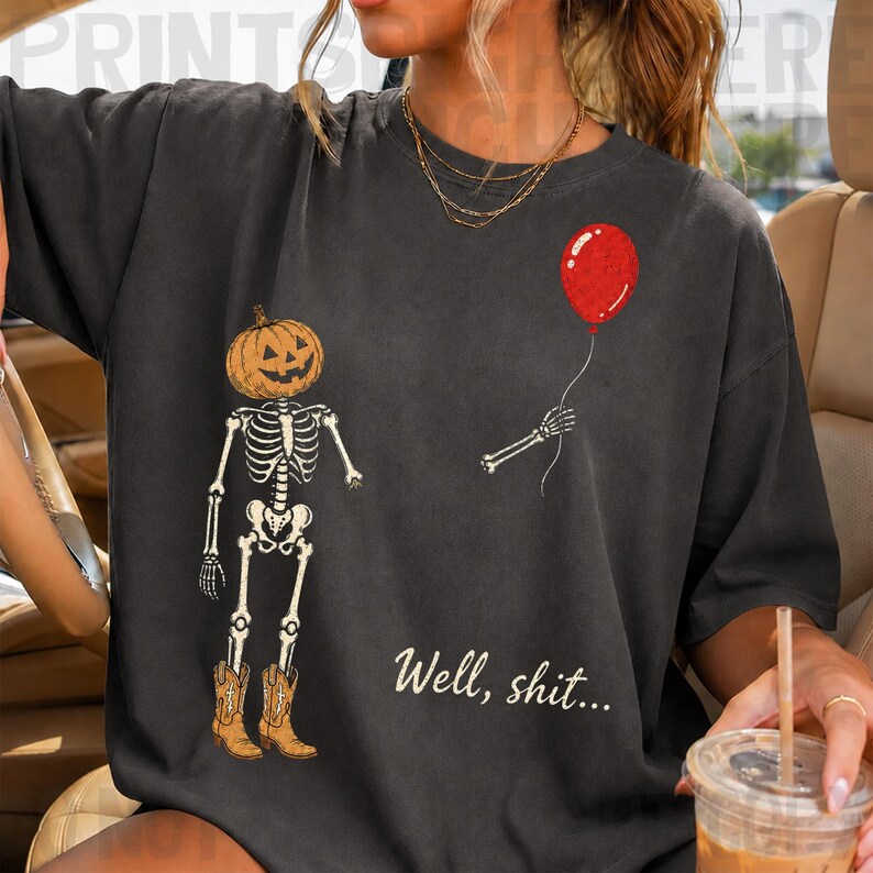 Op de afbeelding: Een antracietgrijs t-shirt met een afbeelding van een skelet met een pompoenhoofd en bruine cowboylaarzen. Het skelet houdt een rode ballon vast in &eacute;&eacute;n hand. De tekst "Well, shit..." staat in een wit cursief lettertype onder de afbeelding.